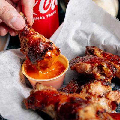A hand dipping a crispy chicken wing into a cup of red sauce beside a pile of wings.