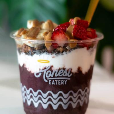 Acai bowl in a clear Honest Eatery cup, Background features green foliage.