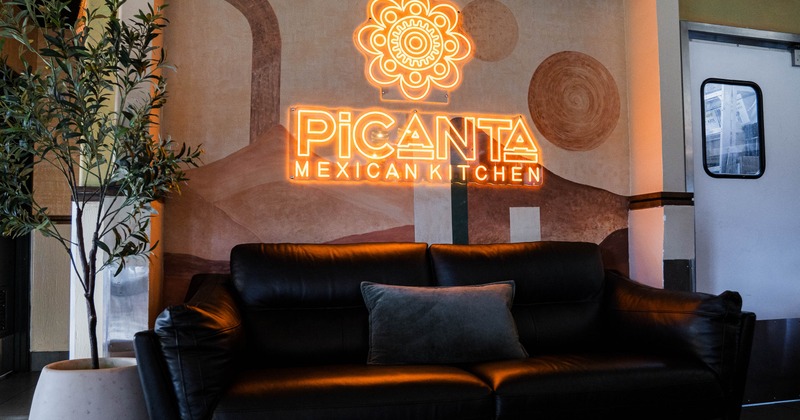 Restaurant lounge area with a black leather sofa, plant and neon sign on a mural wall.