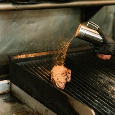 Gloved hand seasoning a steak on a grill grate with a metal shaker.