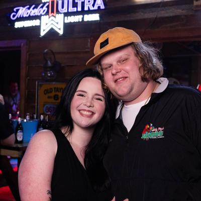 Two  smiling guests pose in the bar with neon signs and soft lighting.