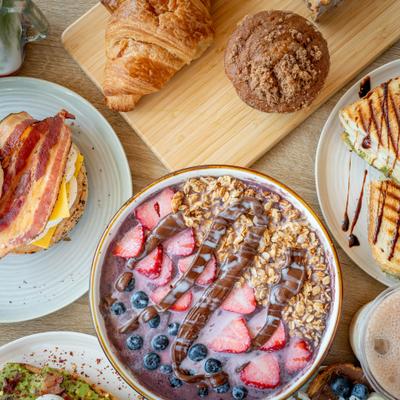 A food spread including an Acai bowl, a breakfast sandwich, baked goods, and beverages.