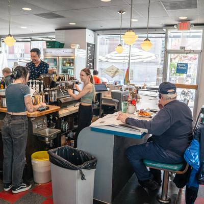 A busy diner scene with staff making drinks behind the counter, A patron dining at the counter.