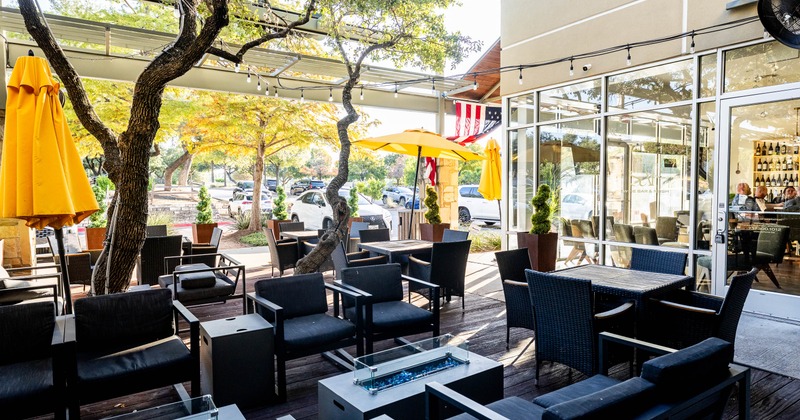 Outdoor patio - dark seating, yellow umbrellas, trees, string lights & a glass-fronted wine bar.
