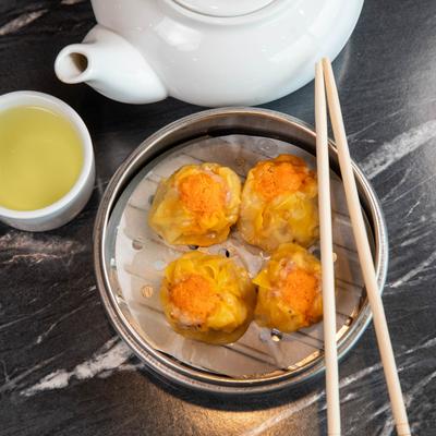 Four pork shumai dumplings topped with roe, accompanied by a teapot and a cup of tea.