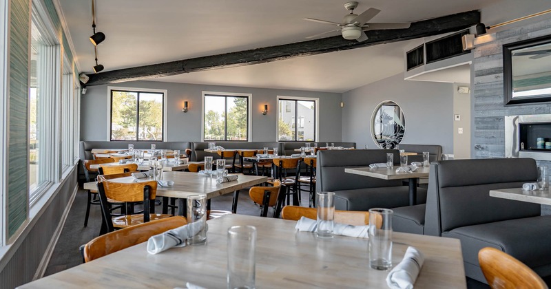 Modern restaurant interior with wooden tables, brown chairs, large windows, and gray booths