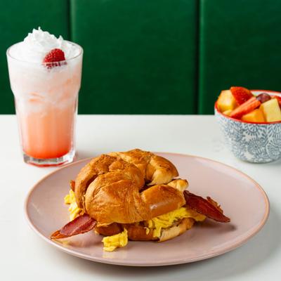 Croissant breakfast sandwich with eggs and bacon served alongside fruit and a drink.