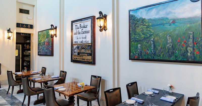 Bright dining room - set tables, black chairs, decorative sconces, framed landscape paintings.