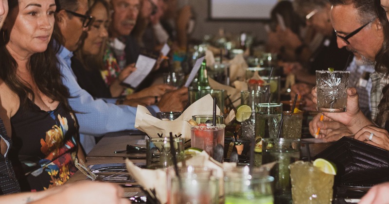 A large group of people sit around a long table, enjoying drinks in a lively, social atmosphere
