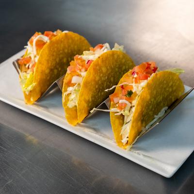 Three hard-shell tacos with cabbage and diced tomatoes on a metal stand.