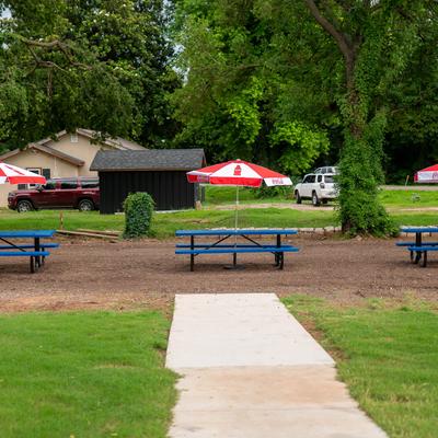 Blue picnic tables with red and white umbrellas in a grassy area next to a paved walkway.