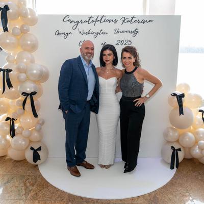 Three people posing before white graduation balloon display.
