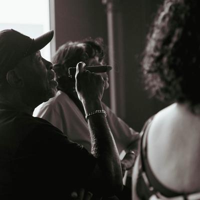 Three people are sitting at a bar counter, one of them is enjoying a cigar.