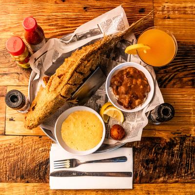 Whole fried fish served with beans, grits, a hushpuppy, and lemon slices on a wooden table.