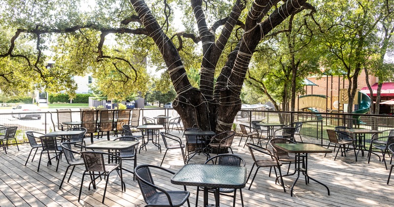 An outdoor restaurant patio features metal tables and chairs shaded by a large tree
