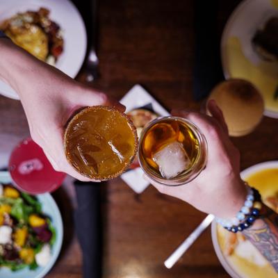 Two hands toasting with cocktails over a table of shared dishes