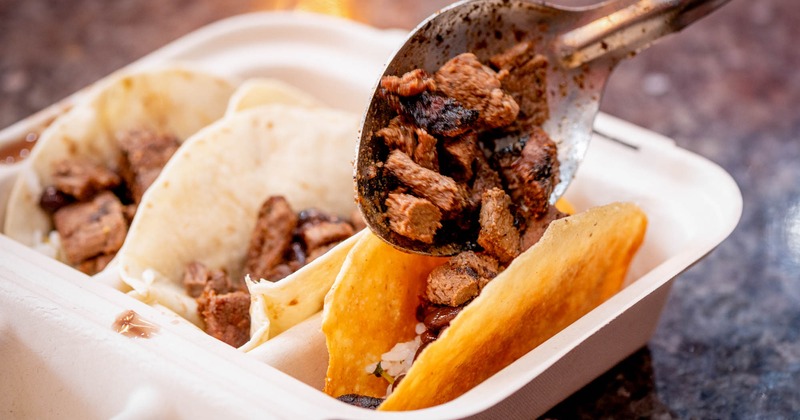 Spoon adding grilled steak to tacos in a takeout container