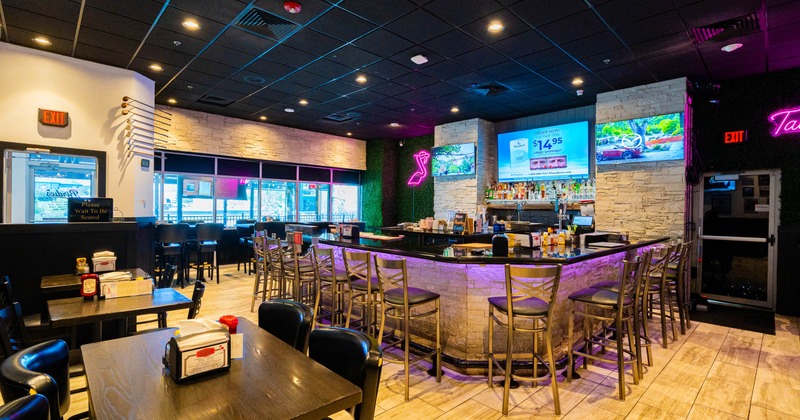 Interior of a brightly lit restaurant with tables, a curved stone-front bar, neon lighting, and wall-mounted screens