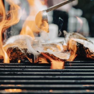 Oysters grilling over flames with sauce poured from a ladle.