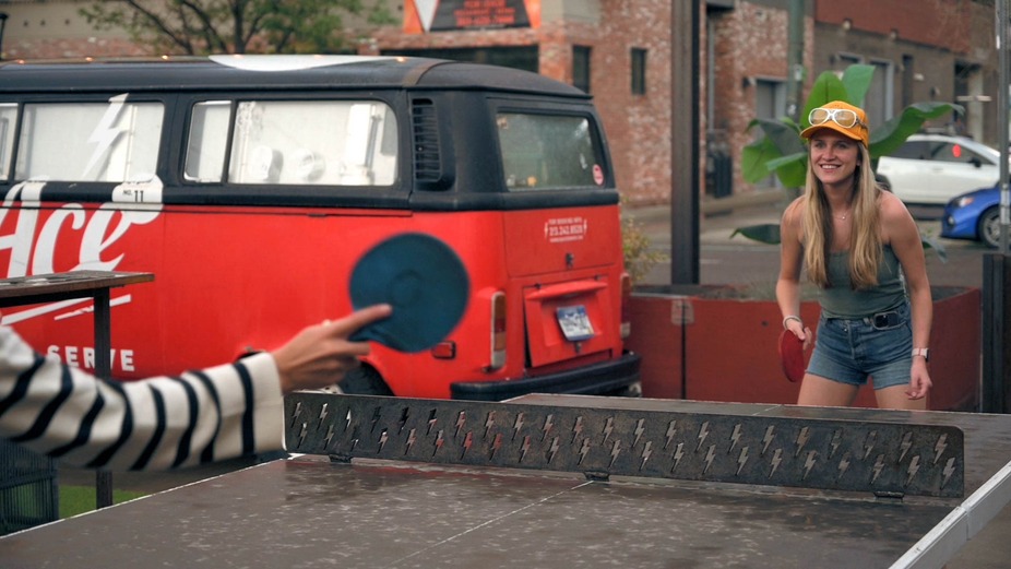 Two people skillfully playing ping pong beside a vibrant red van in an outdoor setting.