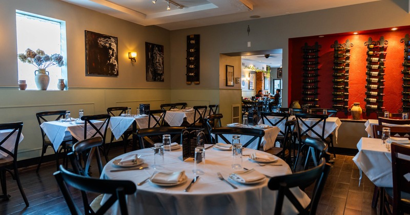 Interior, dining room with white-clothed tables, black chairs, artwork, and a red wine-bottle display wall