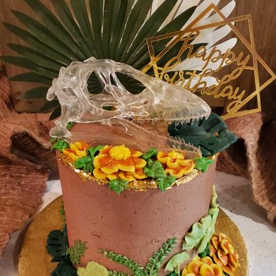 A cake with clear dinosaur skull on top, surrounded by yellow flowers and green leaves.