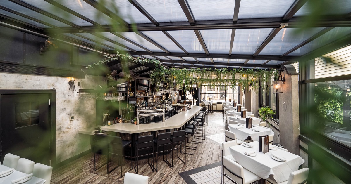 A modern interior with a long bar, white tablecloths, cushioned chairs and hanging greenery