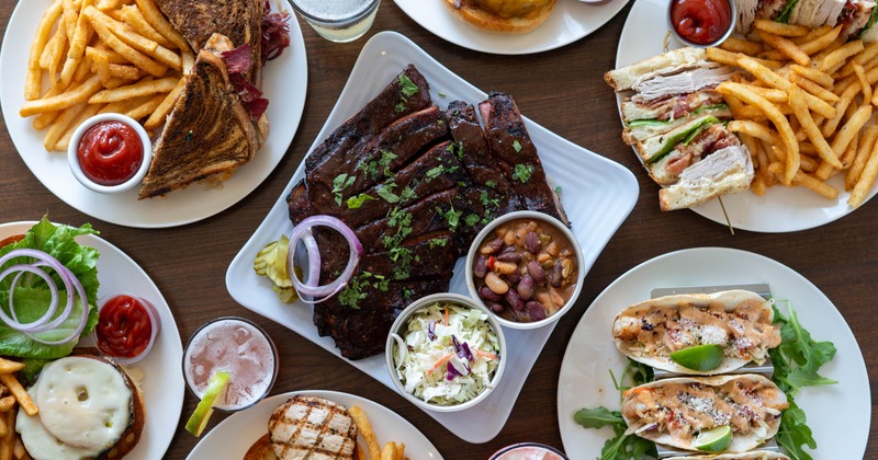 Assorted plates of ribs, sandwiches, burgers, tacos, and fries on a restaurant table