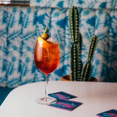A red cocktail on a white table,  set against a blue seating and a potted cactus.
