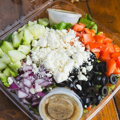Greek salad packed in a clear plastic takeout container with dressing on the side.