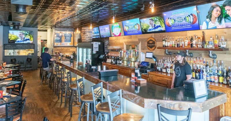 A bartender, a bar stocked with bottles, multiple TVs display sports and media