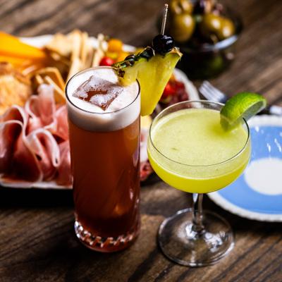 Two colorful cocktails served beside a charcuterie platter on a wooden table.