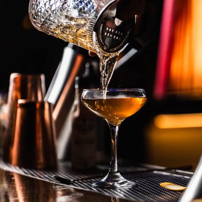 Craft cocktail being poured from a crystal shaker into a coupe glass at a dimly lit bar.