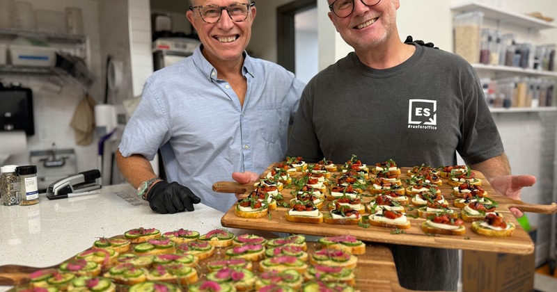 Two people posing, one holding a wooden board of bruschetta
