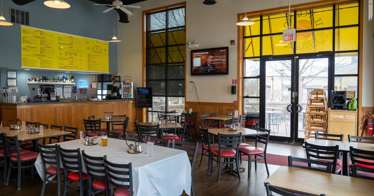 A casual dining area - wooden tables and chairs, a long yellow menu board and a wall-mounted TV.