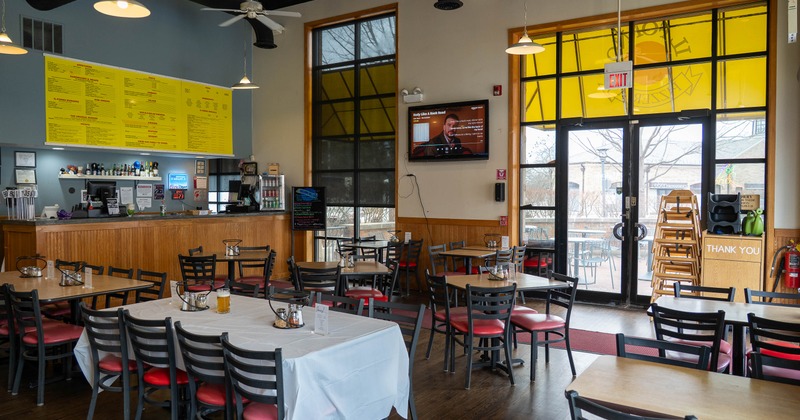 A casual dining area - wooden tables and chairs, a long yellow menu board and a wall-mounted TV.
