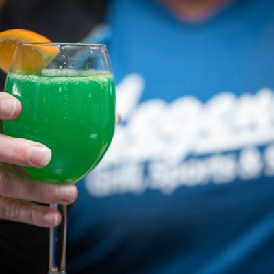 A person holding a glass with a green drink and an orange wedge.