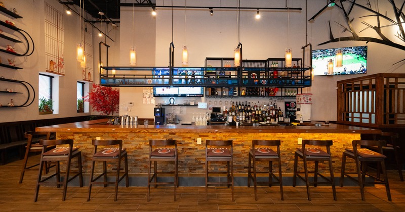 A modern bar with a long wooden counter, bar chairs, mounted shelves with bottles, and flat-screen TVs