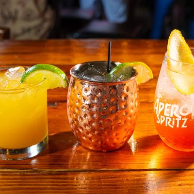 Three colorful cocktails, including a Moscow Mule, sit on a wooden bar.