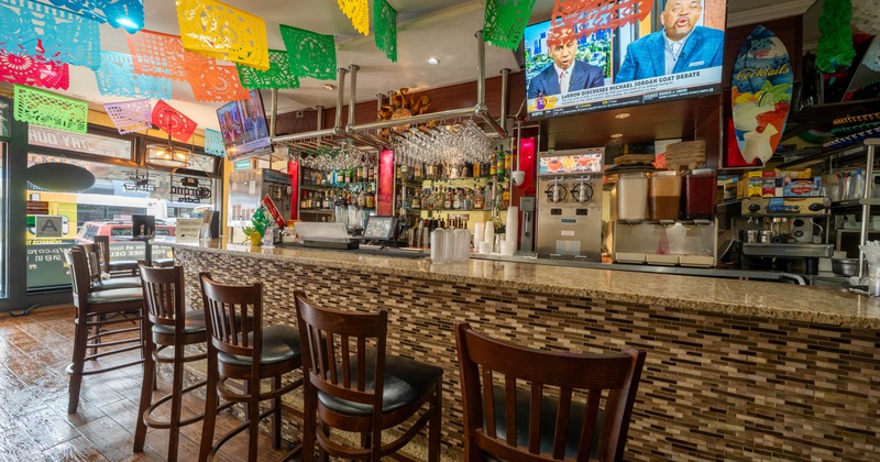 A colorful bar interior with hanging paper banners, wooden stools, and a bar counter