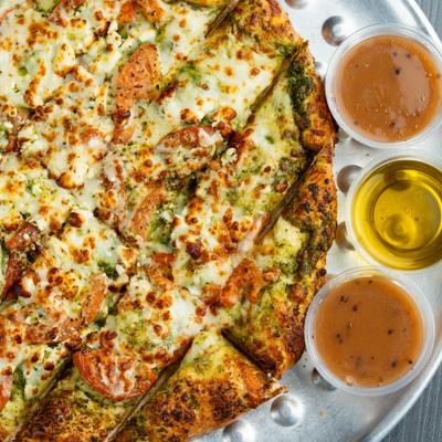 Greek bread featuring a basil pesto base with melted cheese, tomatoes and side of dips.