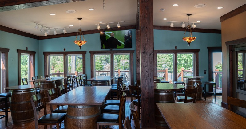 Interior of a rustic restaurant with barrel tables and large windows.