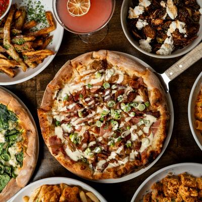 Freshly baked pizza topped with bacon, surrounded by fries, Brussels sprouts, and a cocktail.
