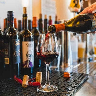 Red wine being poured into a glass on bar counter with assorted wine bottles.