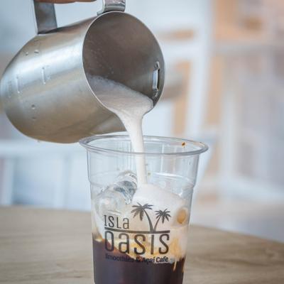 A barista pours steamed milk into a clear plastic cup with an Isla Oasis Smoothies & Acai Cafe logo.