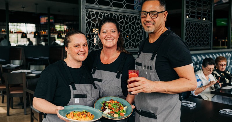 Three staff members hold food plates and a drink at the restaurant