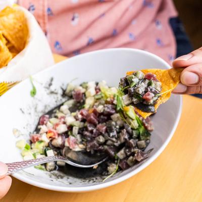 A hand holding a chip topped with black-sauce ceviche over a bowl.