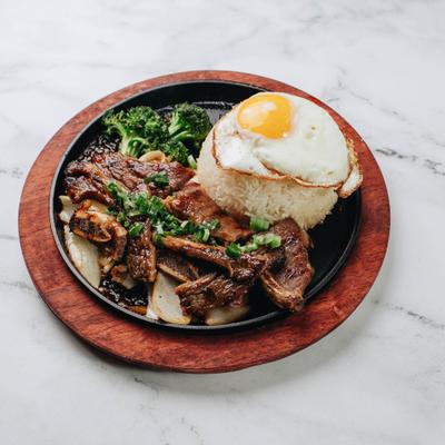 Sizzling Korean beef ribs served on a dark plate with a mound of rice and a fried egg.