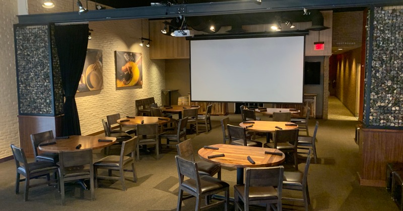 A room with round tables and chairs, and a projector screen