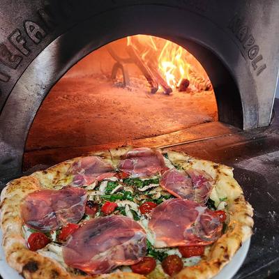 Wood-fired pizza topped with prosciutto and greens in front of a burning oven.